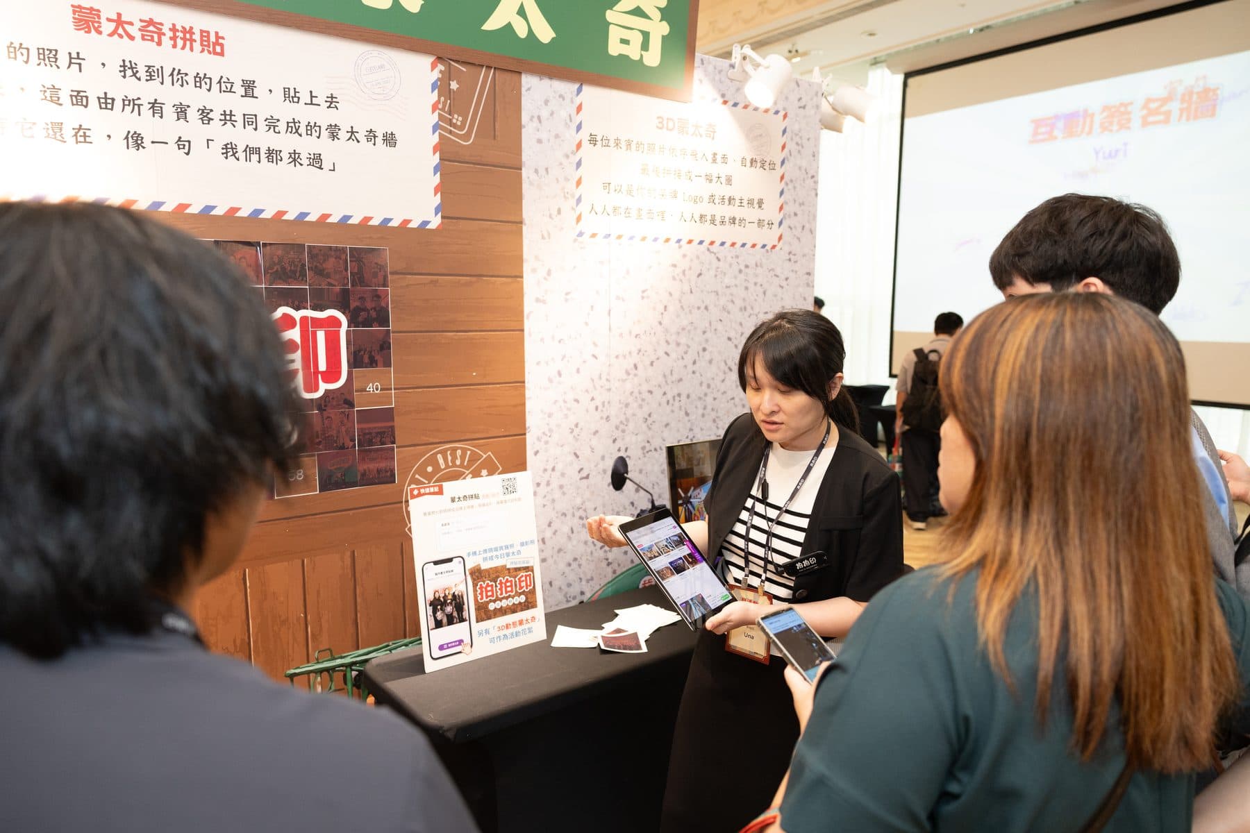 A crew member explaining the photo mosaic wall service beside a wall tiled from many small photographs