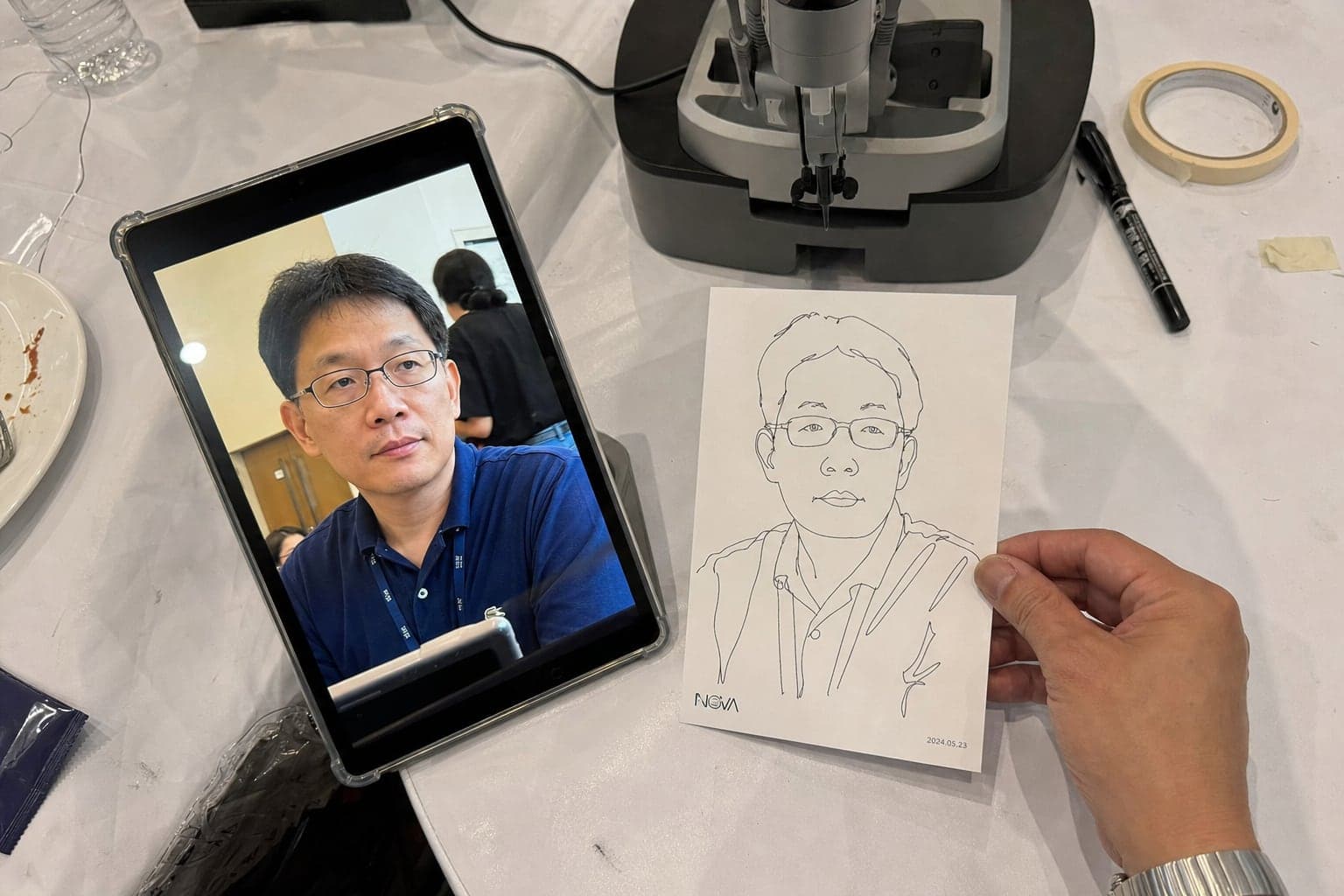 Guest photo on a tablet next to the finished 4×6 line sketch drawn by the robot