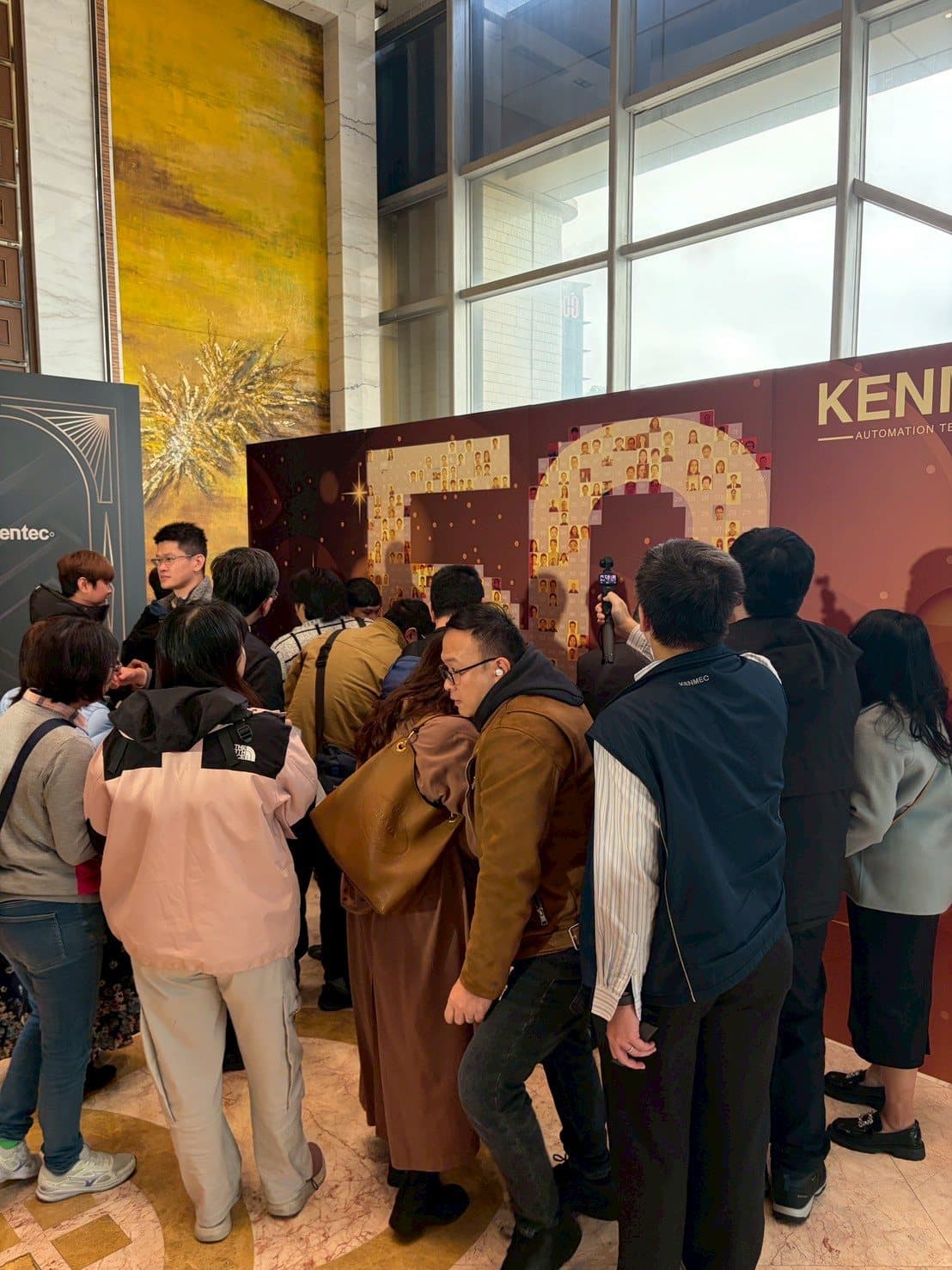 A crowd of guests gathers around the mosaic wall, pasting their photos at a 50th-anniversary celebration