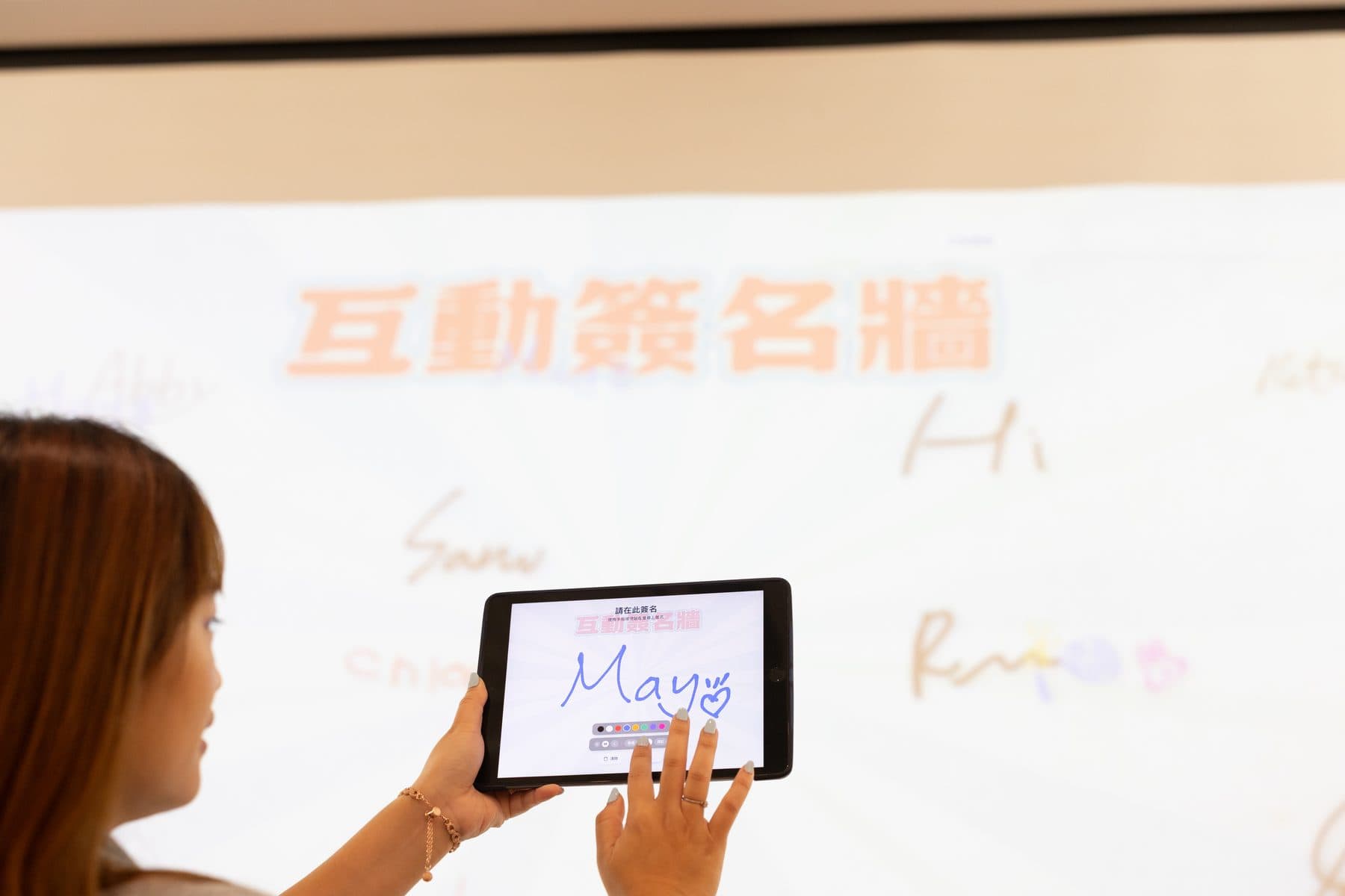 A guest handwriting the name “May” on a tablet, with the signature wall projected on the screen behind